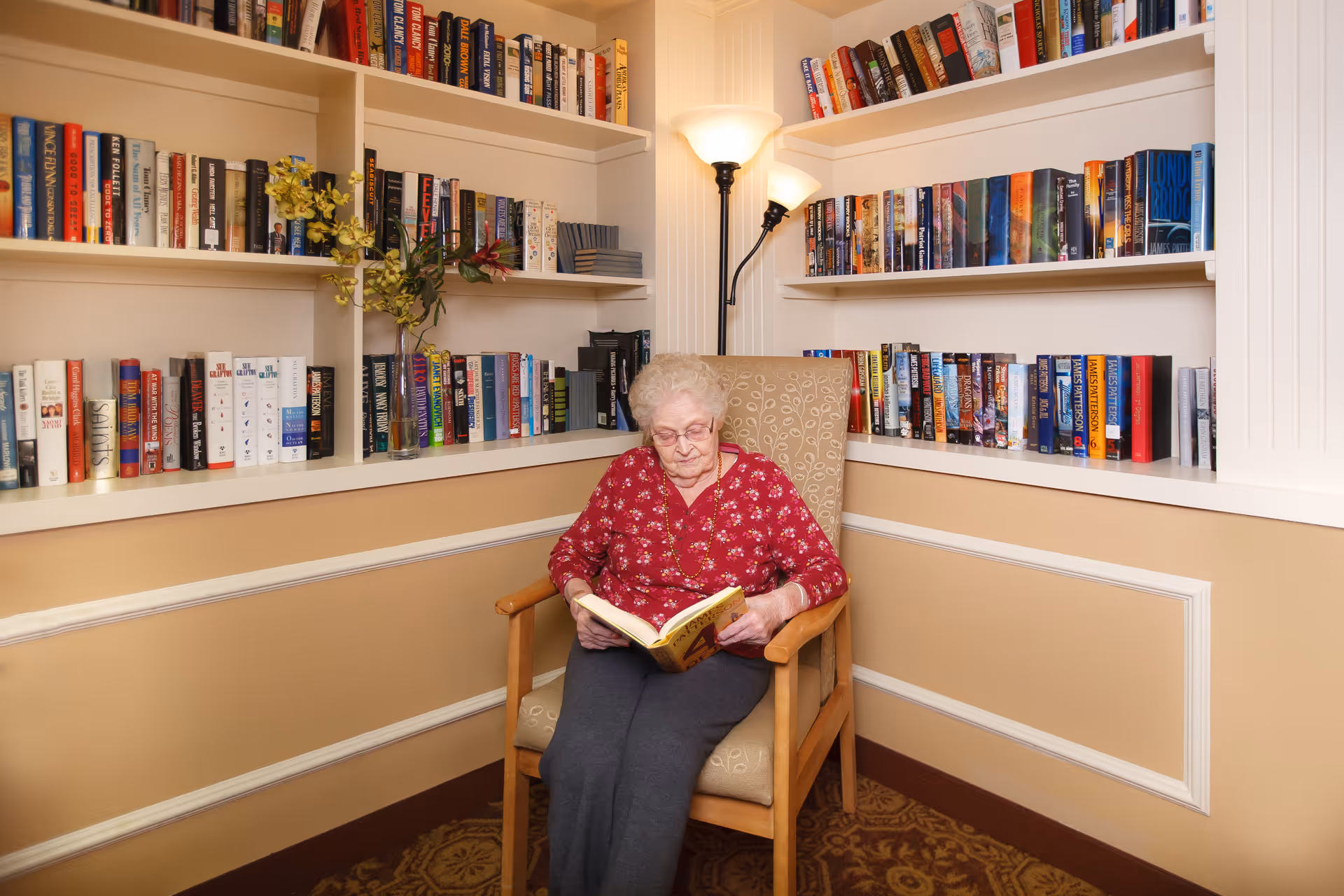 An elderly woman with white hair and glasses is sitting in a beige armchair reading a book. She is wearing a red floral top and dark pants. Behind her are built-in shelves filled with numerous books and a floor lamp with two lights.