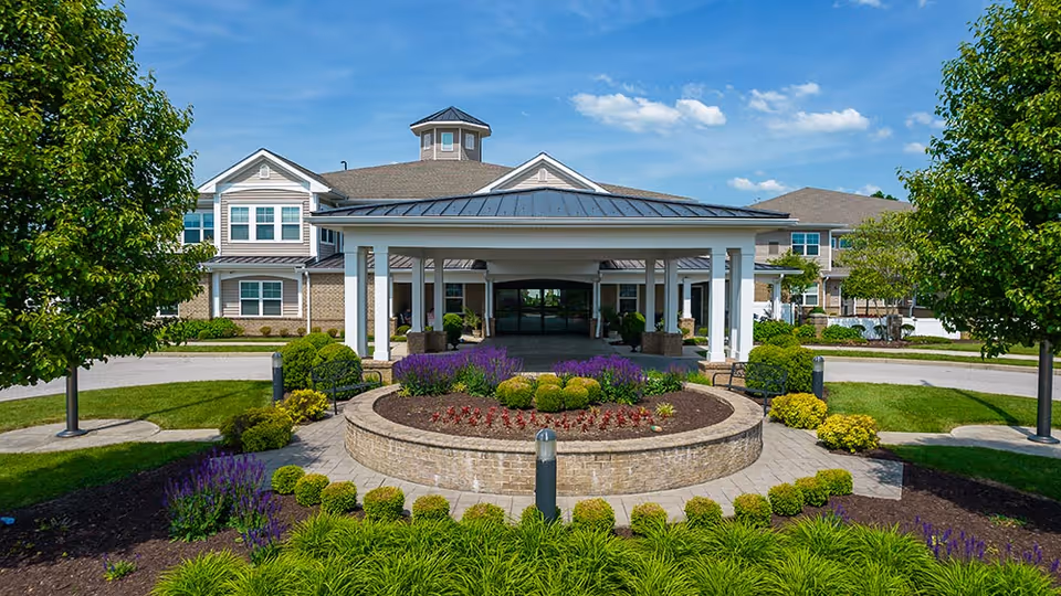 Front exterior view of English Meadows facility with a covered entrance, landscaped garden featuring bushes and flowers, and a clear blue sky.