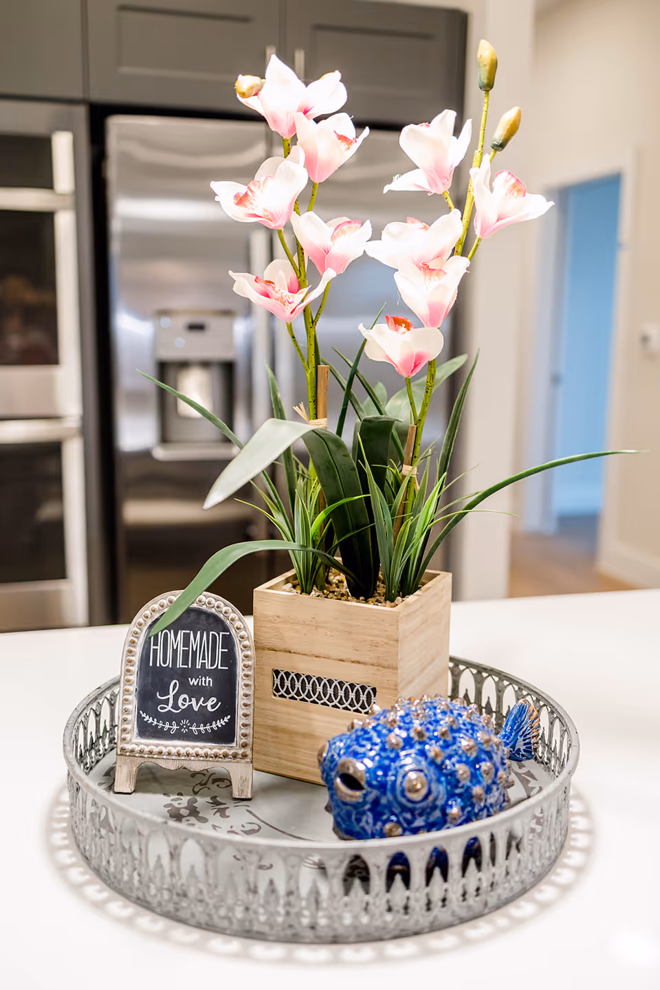 A decorative tray on a kitchen counter holding a wooden planter with pink and white artificial flowers, a small sign that reads 'HOMEMADE with Love,' and a blue ceramic fish figurine. In the background, there is a stainless steel refrigerator and an oven.