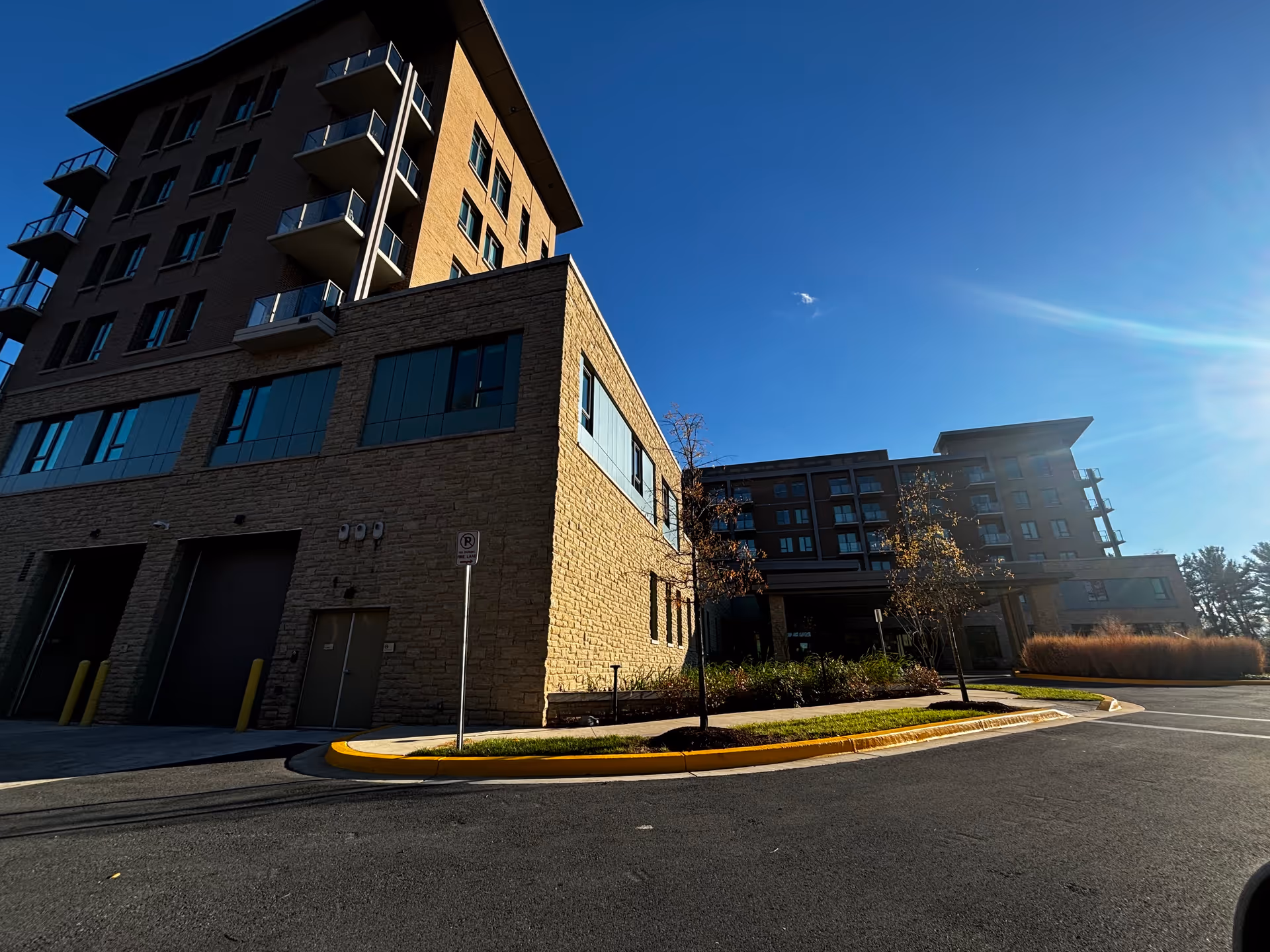 Sunlit multi-story senior living building with balconies, entrance driveway, and landscaping under a clear blue sky.