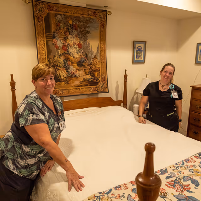 Two women in uniform standing on either side of a made bed in a bedroom with a wooden headboard, a floral tapestry hanging on the wall above the bed, and framed pictures on the walls.