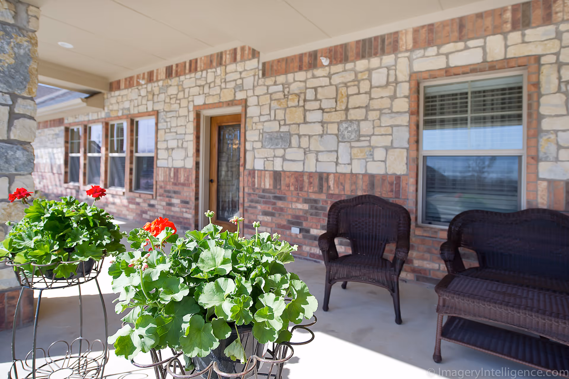 A covered outdoor patio area with stone and brick walls, featuring two dark wicker chairs and a matching wicker loveseat. In the foreground, there are green plants with red flowers in metal plant stands. A door with a glass panel and several windows are visible along the wall.