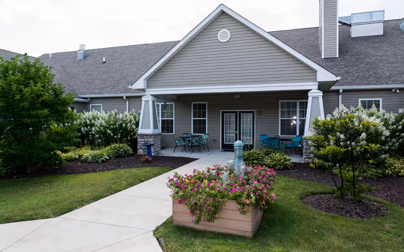 Exterior view of a single-story senior living facility building with a covered entrance, patio furniture, and landscaped garden beds with flowers and shrubs along a concrete walkway.