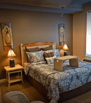Cozy bedroom with a made bed featuring patterned bedding, bedside tables and lamps, wall art, and a window air conditioner.