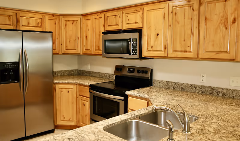 A kitchen with wooden cabinets, granite countertops, a stainless steel refrigerator, a built-in microwave above a black and stainless steel electric stove, and a double sink with a modern faucet.