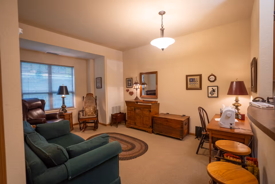 Cozy living room with a green sofa, armchairs, wooden cabinets and desk (with a sewing machine), lamps, and warm overhead lighting.