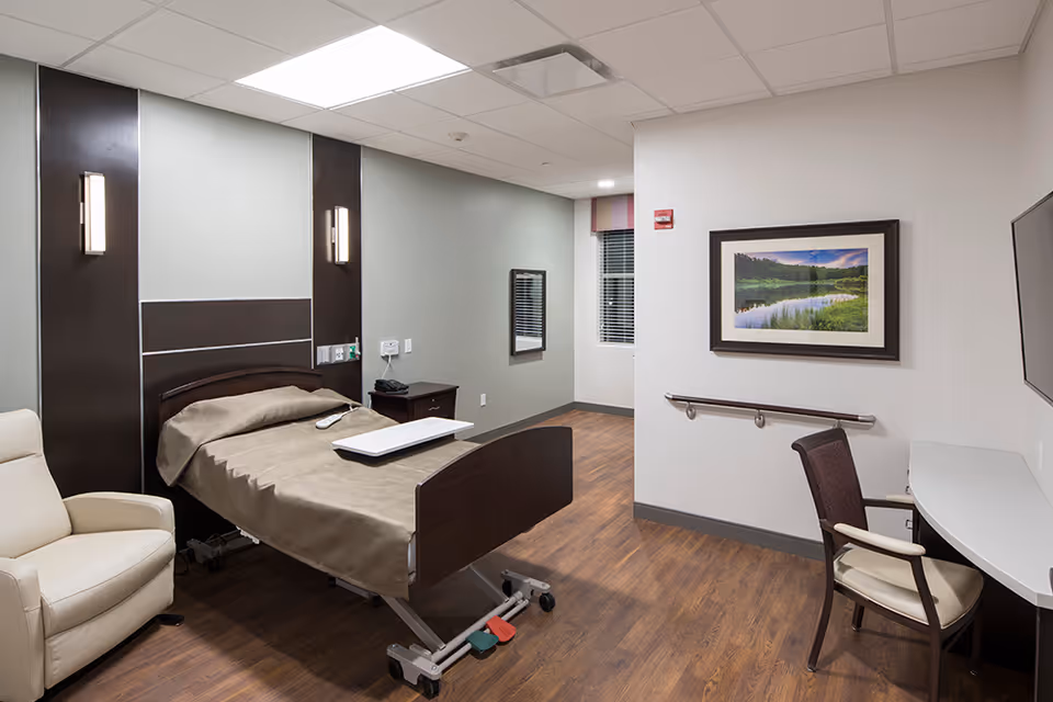 A clean and modern bedroom in a senior living facility featuring a hospital-style adjustable bed with beige bedding, a beige recliner chair, a wooden nightstand, a wall-mounted flat screen TV, a desk with a chair, and a framed landscape picture on the wall. The room has wood flooring and neutral-colored walls with two vertical wall lights beside the bed.