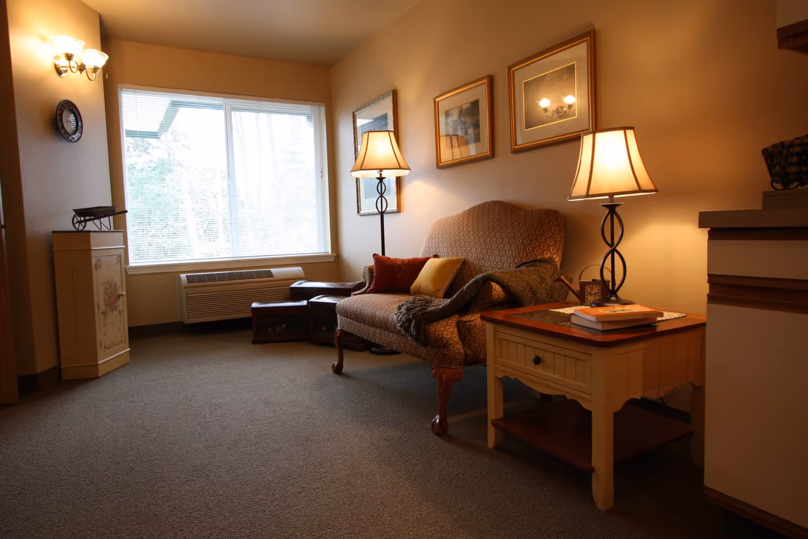 A cozy living room area with a patterned loveseat adorned with two pillows and a throw blanket. There are two table lamps with warm lighting, one on a side table next to the loveseat and another standing lamp behind it. The walls have framed pictures and a mirror, and a large window with blinds lets in natural light. The room has carpeted flooring and a small cabinet with decorative items on the left side.