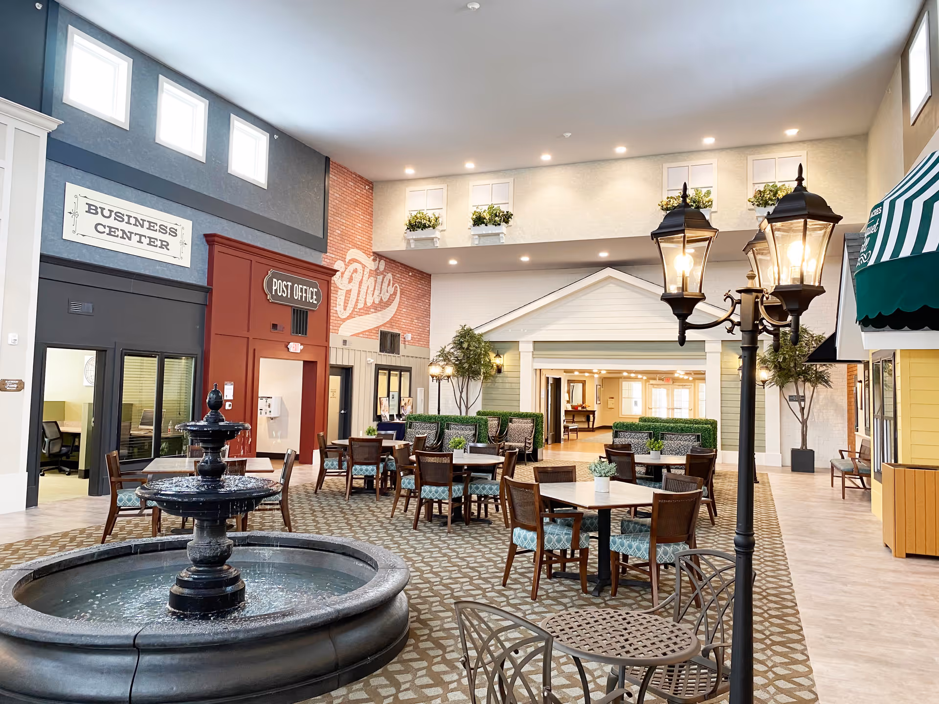 Indoor common area designed like a small town square with a black water fountain in the foreground, several tables and chairs with patterned cushions, a vintage-style street lamp, and storefront facades labeled Business Center and Post Office. The walls have decorative windows with flower boxes and a mural that says Ohio.