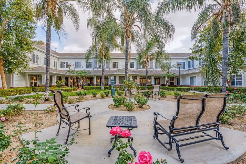 Outdoor courtyard area of a senior living facility with palm trees, rose bushes, and several metal chairs and benches arranged around a small table on a concrete patio. The two-story building with white siding and multiple windows surrounds the courtyard.