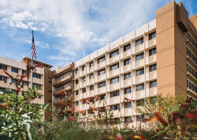 Exterior view of a multi-story beige and white building with many windows, an American flag on a pole, and greenery with flowers in the foreground under a partly cloudy sky.