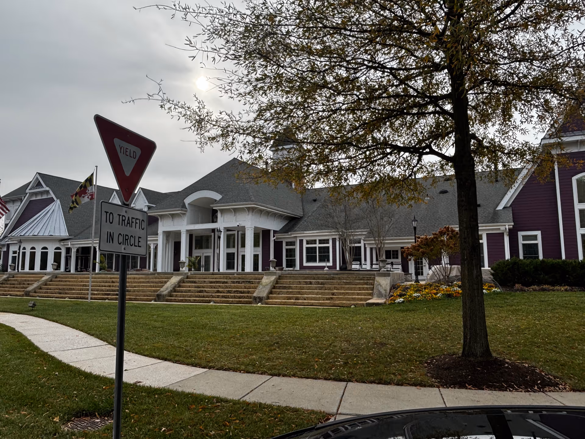 Exterior view of a large building with a gray roof and purple siding, featuring white columns and multiple windows. There are steps leading up to the entrance, a tree in the foreground, and a yield traffic sign with an additional sign below it that reads 'TO TRAFFIC IN CIRCLE'. The sky is overcast.