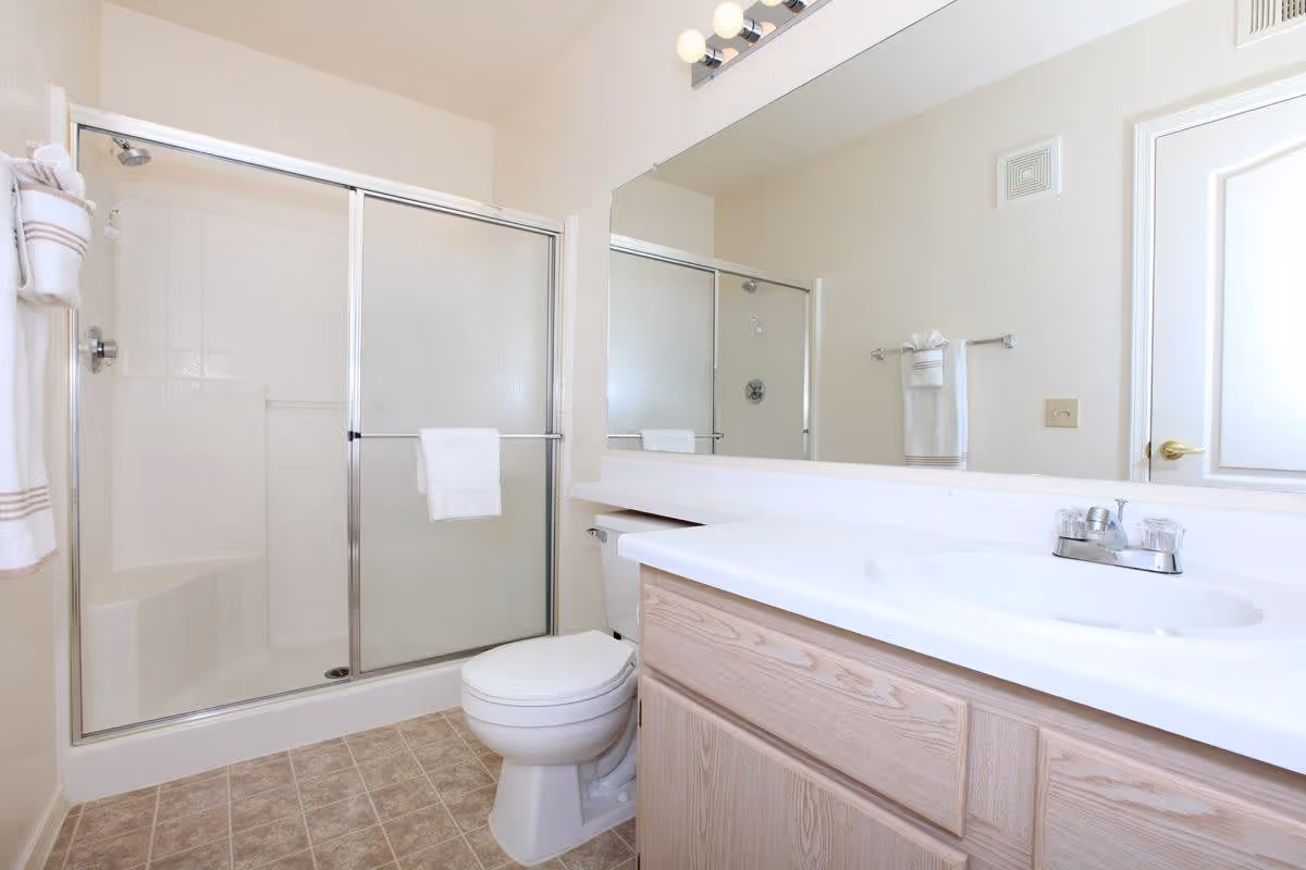 A clean bathroom with a glass-enclosed shower, a white toilet, a long white countertop with a sink, wooden cabinets underneath, a large mirror above the sink, and white towels hanging on towel racks.