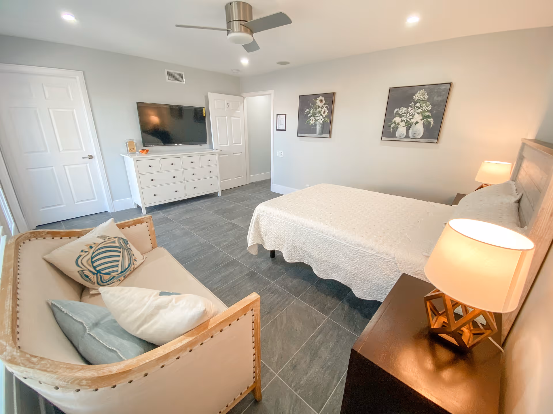 A bright and clean senior care bedroom featuring a bed with a white quilt, two bedside tables with lamps, a small couch with decorative pillows, a white dresser with a flat-screen TV mounted above it, and two floral paintings on the wall.