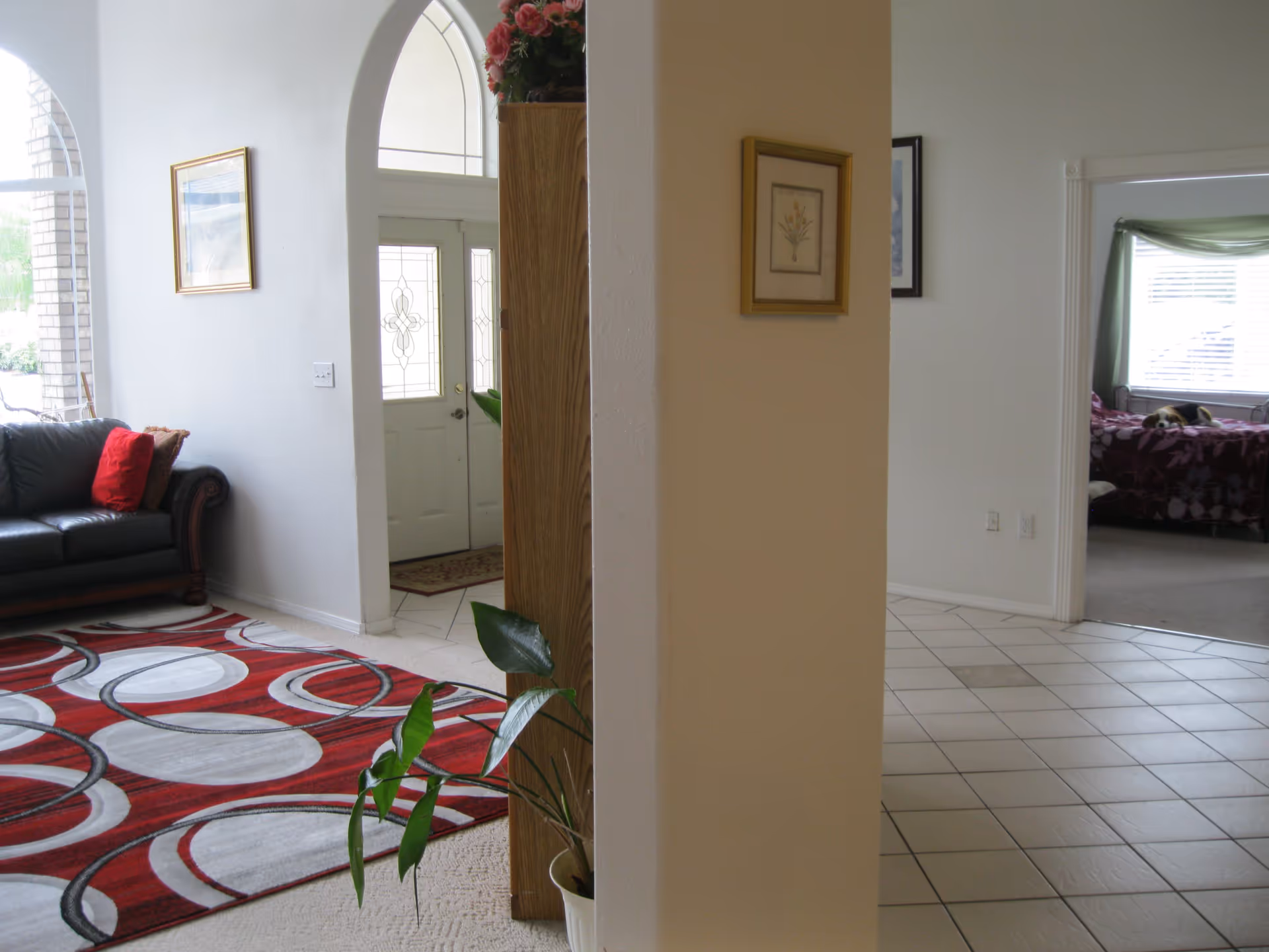 Interior view showing a living room with a leather sofa, red-and-white rug, an arched entryway, and a bedroom visible through a doorway.