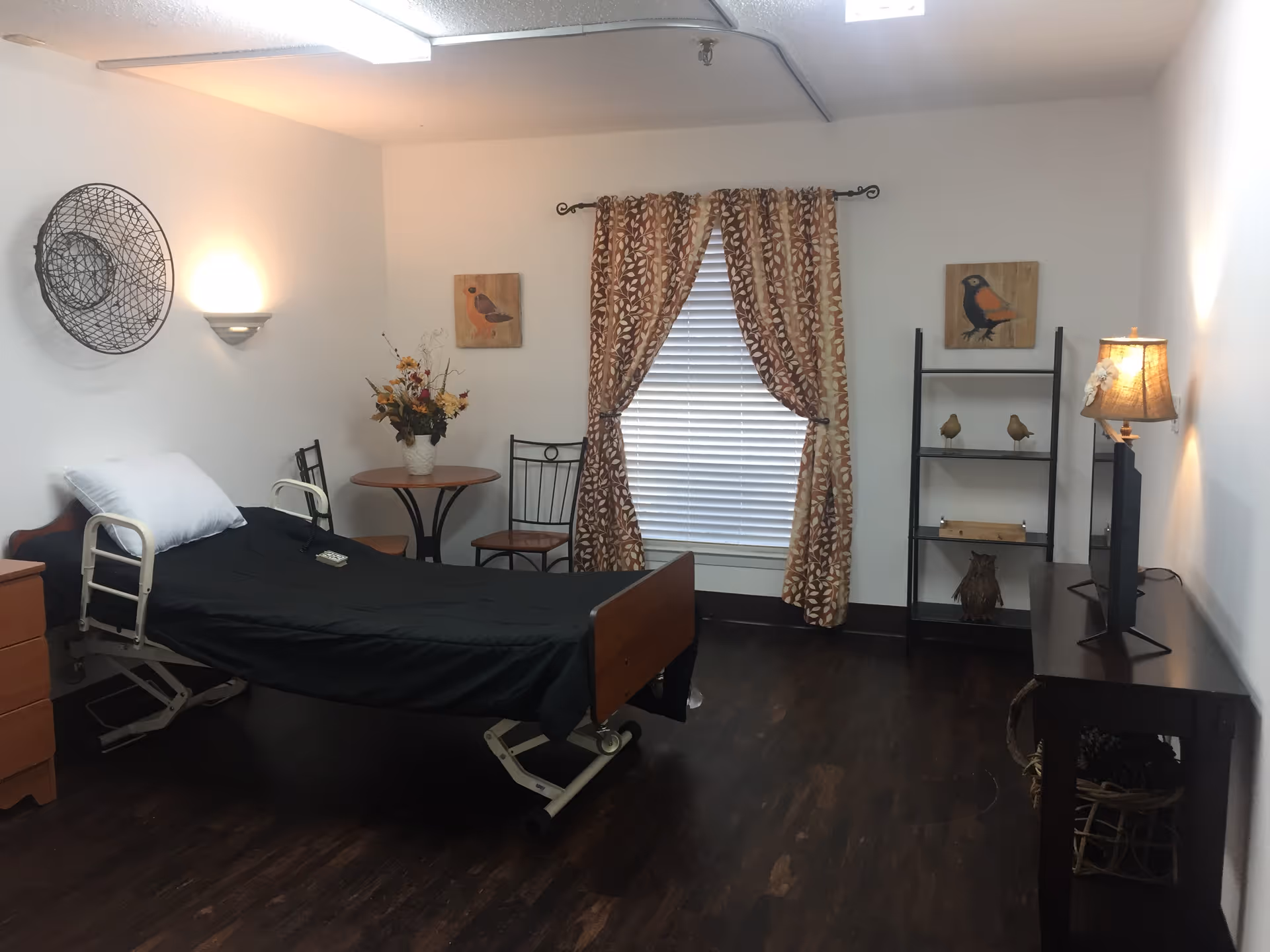 A simple, clean room with a hospital-style bed covered in a black blanket and a white pillow. The room has dark wood flooring, a small round table with two chairs, a window with patterned curtains, a wall-mounted light fixture, and decorative wall art featuring birds. There is a black shelving unit with small bird figurines and a lamp on a dark wooden table with a TV on it.