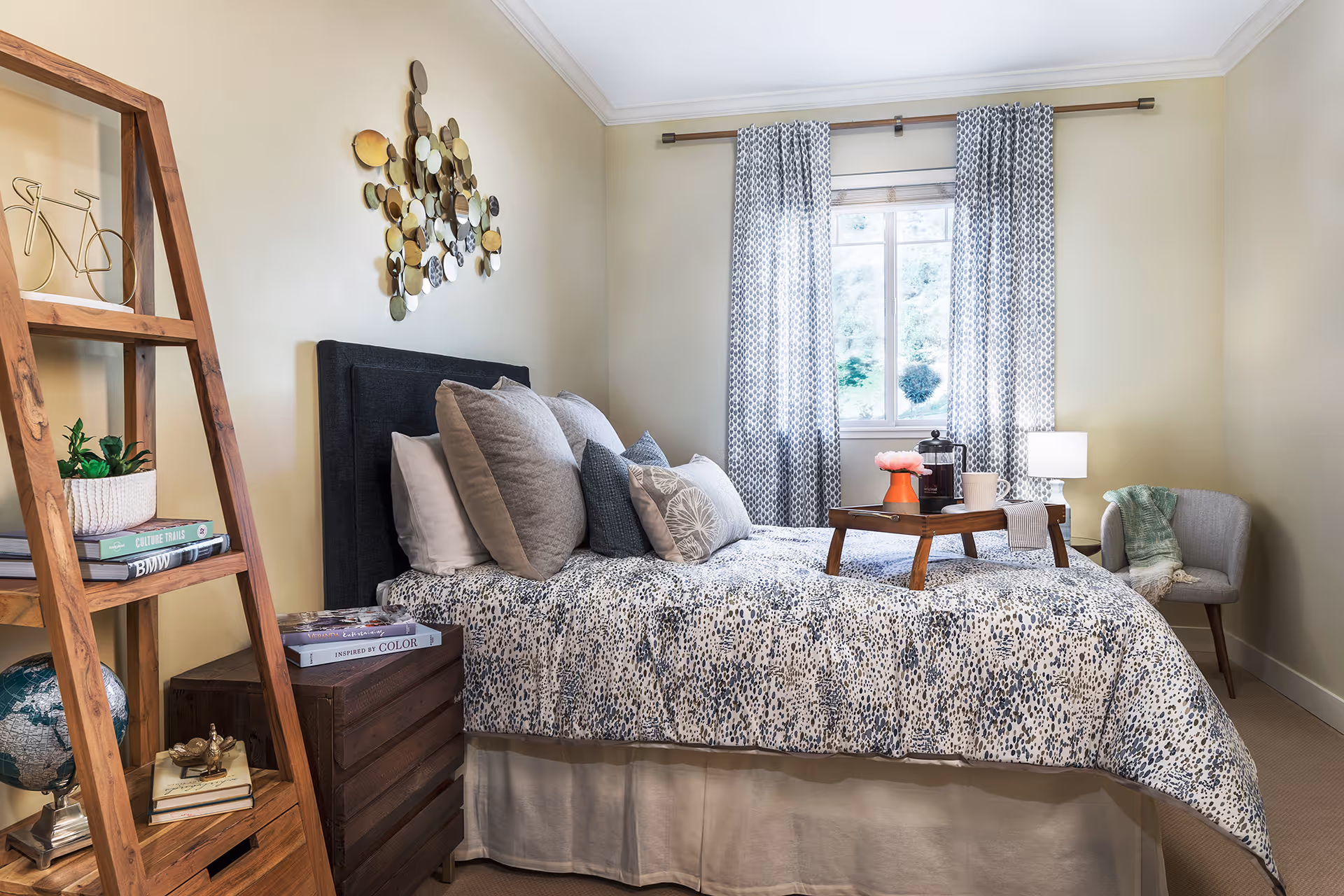 A cozy bedroom with a bed covered in a patterned comforter and multiple pillows. A wooden tray with a coffee pot, cup, and a small vase with flowers sits on the bed. There is a window with patterned curtains behind the bed, a small gray armchair with a green throw blanket to the right, and a wooden ladder-style shelf with books, a globe, and decorative items to the left. The walls are painted a light beige color.