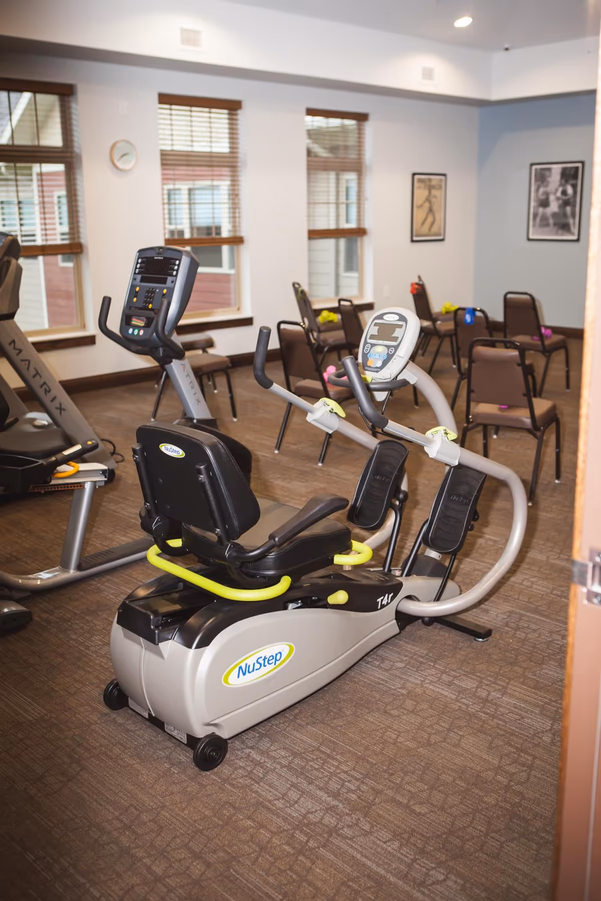 Exercise room with stationary fitness equipment including a NuStep recumbent cross trainer and a Matrix treadmill. Several chairs are arranged in the background near windows with wooden blinds. The room has framed pictures on the walls and carpeted flooring.