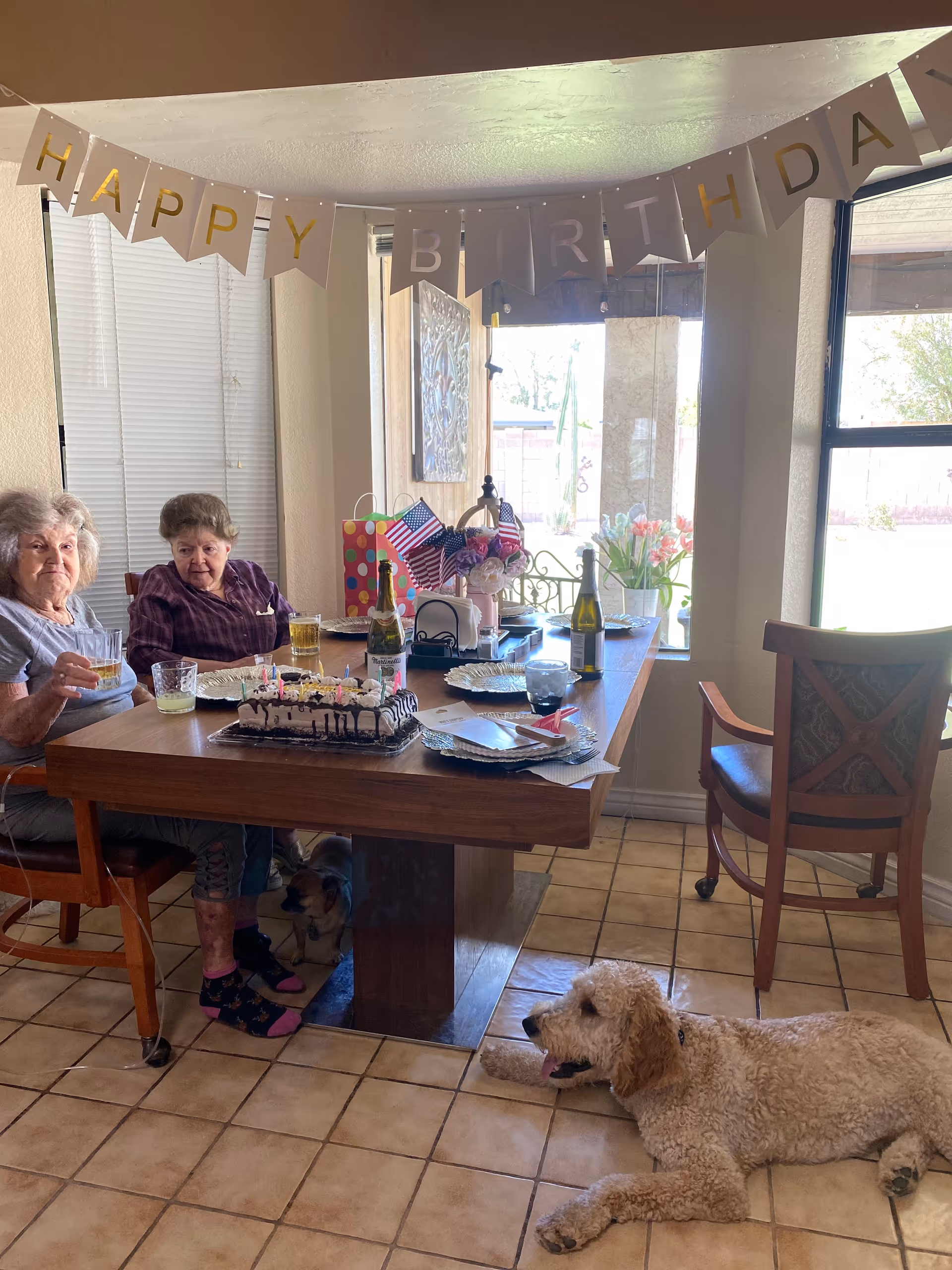 Two older women sit at a dining table decorated for a birthday with cake, gifts and drinks while a dog rests on the tile floor.