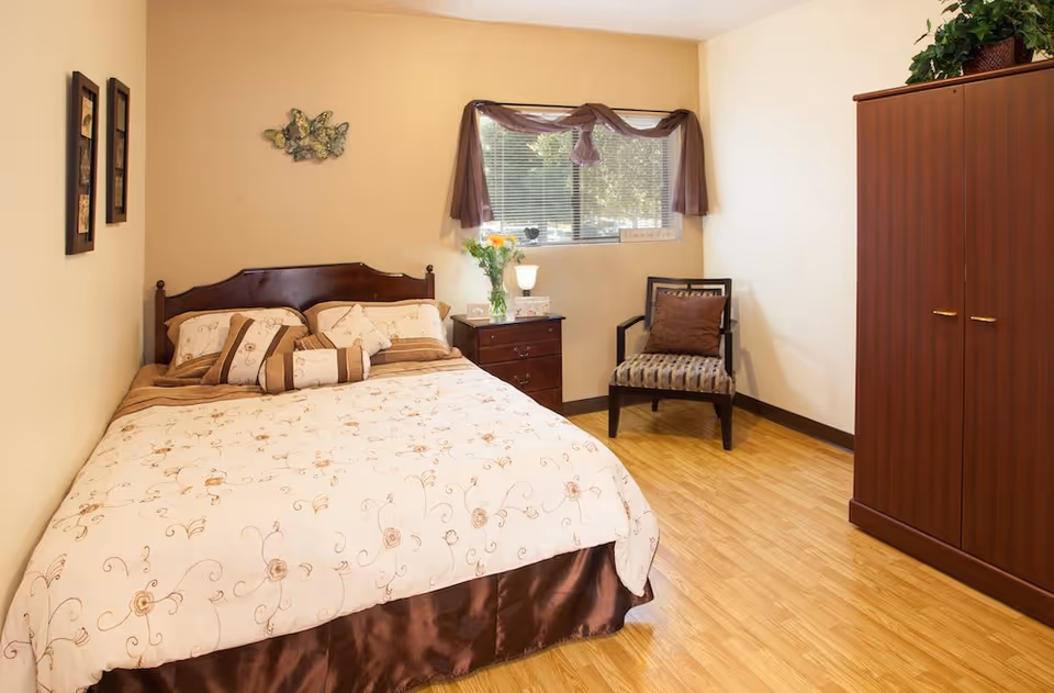 A cozy bedroom with a double bed covered in a cream and brown floral patterned comforter. There is a wooden nightstand with a lamp and a vase of flowers next to the bed. A window with brown curtains lets in natural light. A cushioned chair with a brown pillow and a wooden wardrobe are also visible. The floor is wooden and the walls are painted light beige.