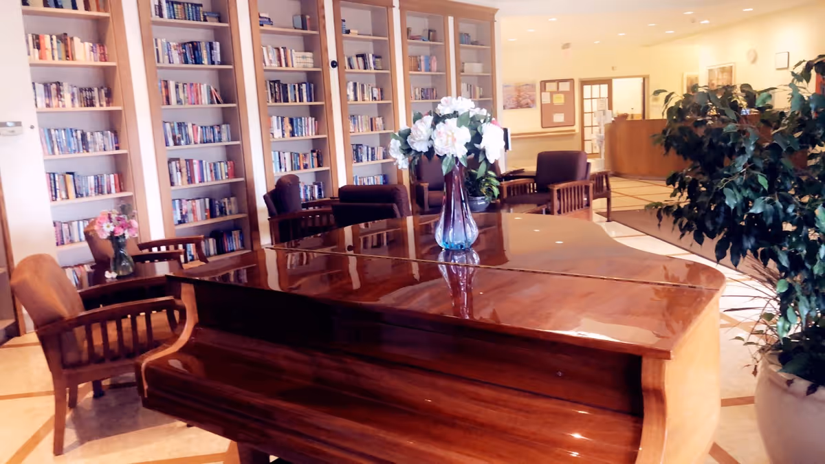 A polished grand piano sits in a library-like lounge with bookshelves and plants.