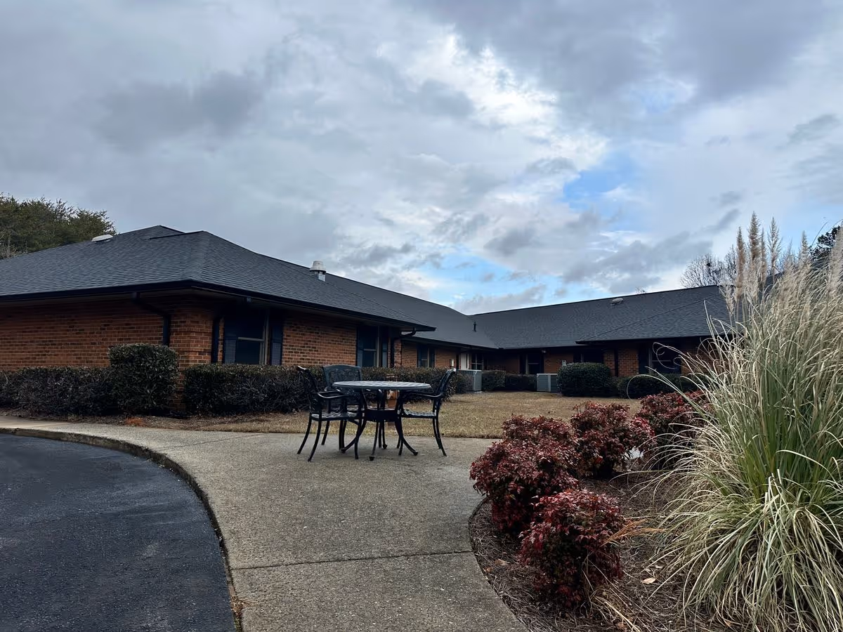 Outdoor courtyard of a single-story brick senior living building with a patio table and chairs, landscaped shrubs, and a cloudy sky.