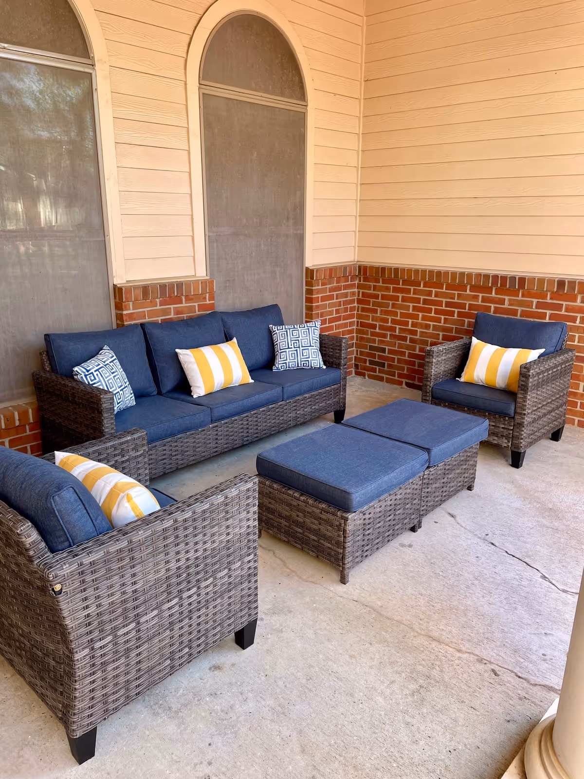 A covered outdoor patio with wicker seating, blue cushions, and yellow-striped pillows against a brick and siding wall.