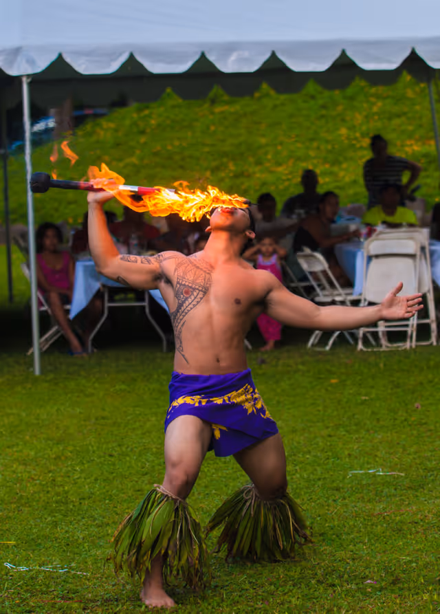 A shirtless man with tribal tattoos performs a fire-eating act outdoors on grass. He wears a purple cloth with yellow patterns around his waist and green leaf decorations around his lower legs. In the background, people sit under a white canopy tent watching the performance.