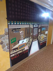 A mural painted on an interior wall depicting a storefront scene with signs for Florsheim shoes, Jets, and Vogel's Shoe Store. The mural includes displays of shoes and a window with text, set against a yellow wall with stone accents and a carpeted floor.