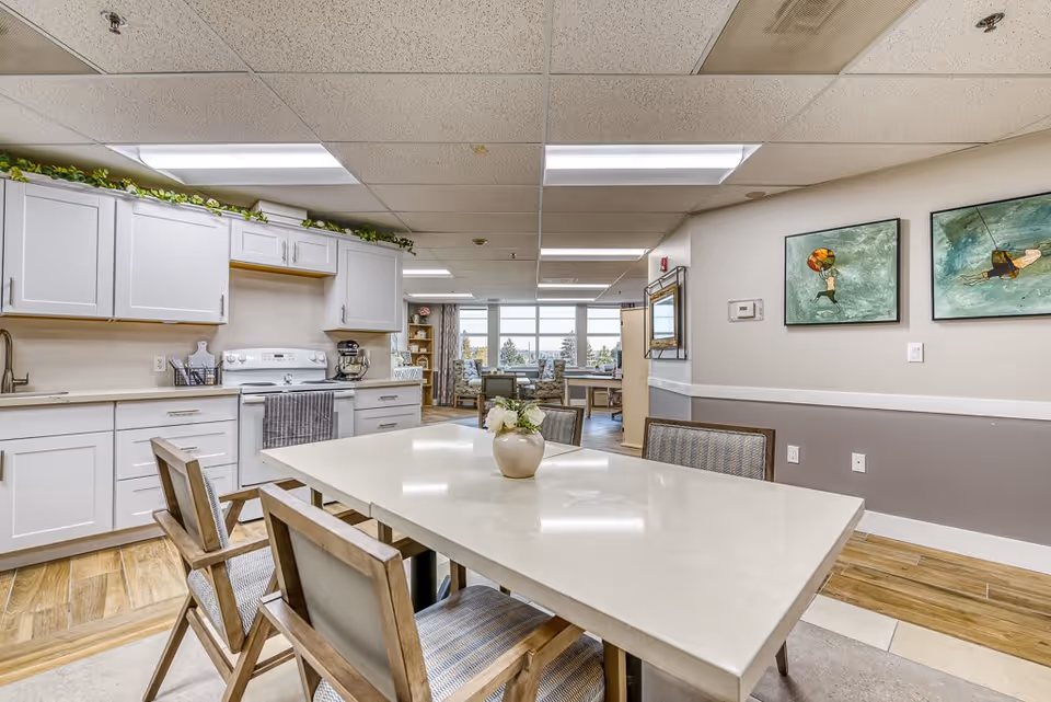 A bright and clean kitchen and dining area in a senior living facility. The kitchen features white cabinets, a white stove, and a countertop with a mixer. In the foreground, there is a dining table with four wooden chairs and a small vase with flowers on the table. The room has wood flooring and fluorescent ceiling lights. On the right wall, there are two framed paintings and a thermostat. In the background, there is a seating area with large windows letting in natural light.