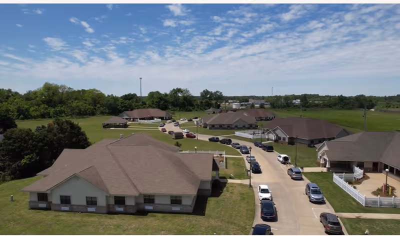 Aerial view of a senior living facility with multiple single-story buildings with brown roofs, surrounded by green lawns and trees. A curved driveway runs through the property with several parked cars along it under a partly cloudy sky.