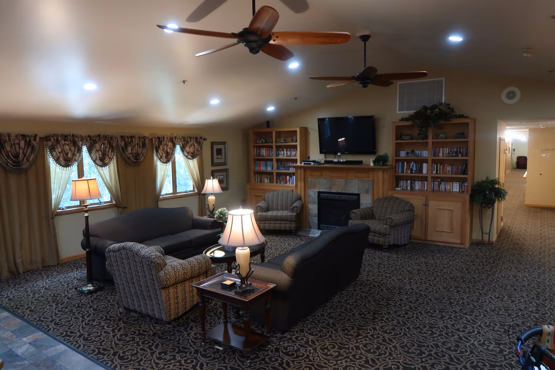 A cozy senior living facility common area with patterned carpet, two ceiling fans, and multiple seating options including two sofas and two armchairs arranged around a coffee table with a lamp. There are two floor lamps near the windows with draped curtains. A fireplace with a mounted TV above it is flanked by two wooden bookshelves filled with books and decorative plants. A hallway is visible to the right.