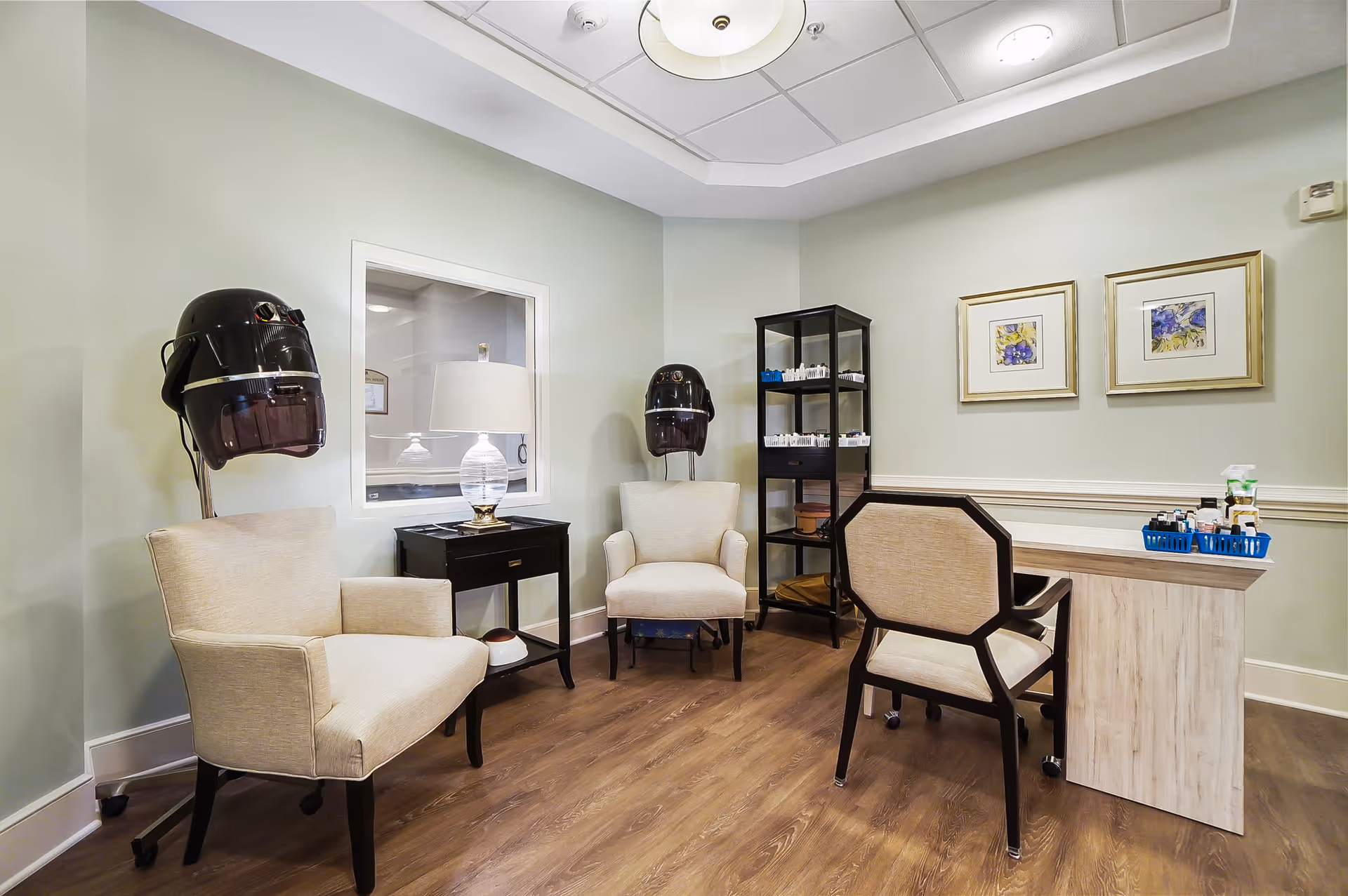 Interior salon room with two styling chairs under hair dryers, a manicure table, shelving with supplies, and framed artwork.