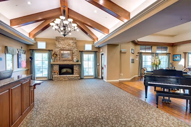 Spacious senior living facility common area with a high vaulted ceiling featuring wooden beams and a chandelier. A stone fireplace with a framed painting above it is centered between two large windows with curtains. To the right, there is a black grand piano on a wooden floor next to a dining area with chairs and a table decorated with flowers. The left side shows a wooden reception desk with a computer monitor.