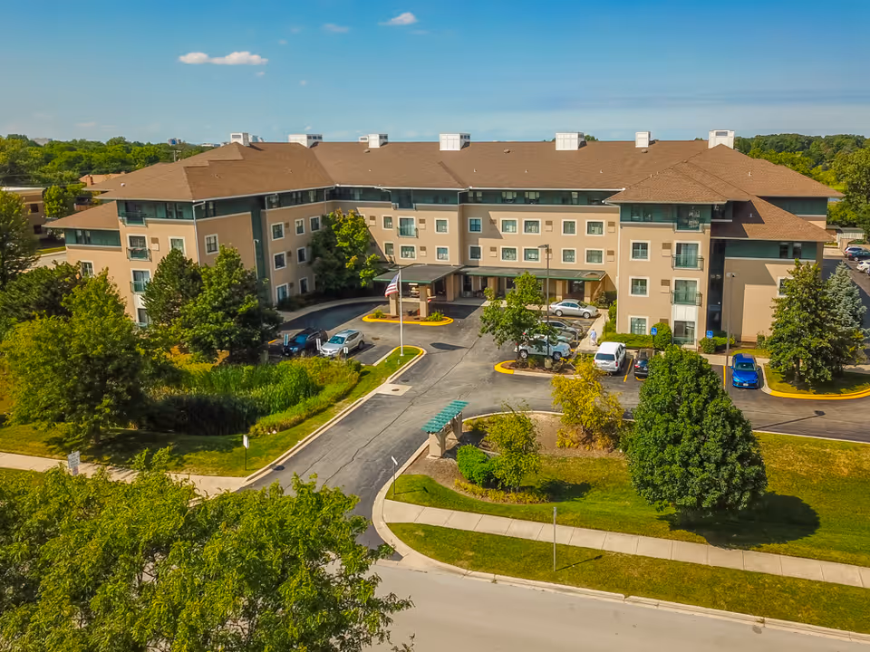 Aerial view of Cordia Senior Residence - Westmont, a multi-story senior living facility with a brown roof and beige exterior walls. The building is surrounded by green trees, landscaped lawns, and a parking area with several cars. The entrance features a covered drop-off area with an American flag on a flagpole nearby.