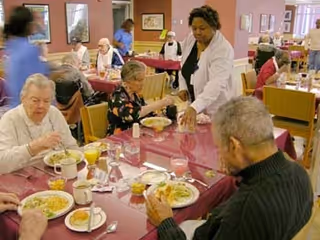 Several elderly residents seated at tables covered with red tablecloths in a dining room, eating meals. A staff member in a white coat is serving food to one of the residents. Other staff members in blue uniforms are visible in the background assisting residents.