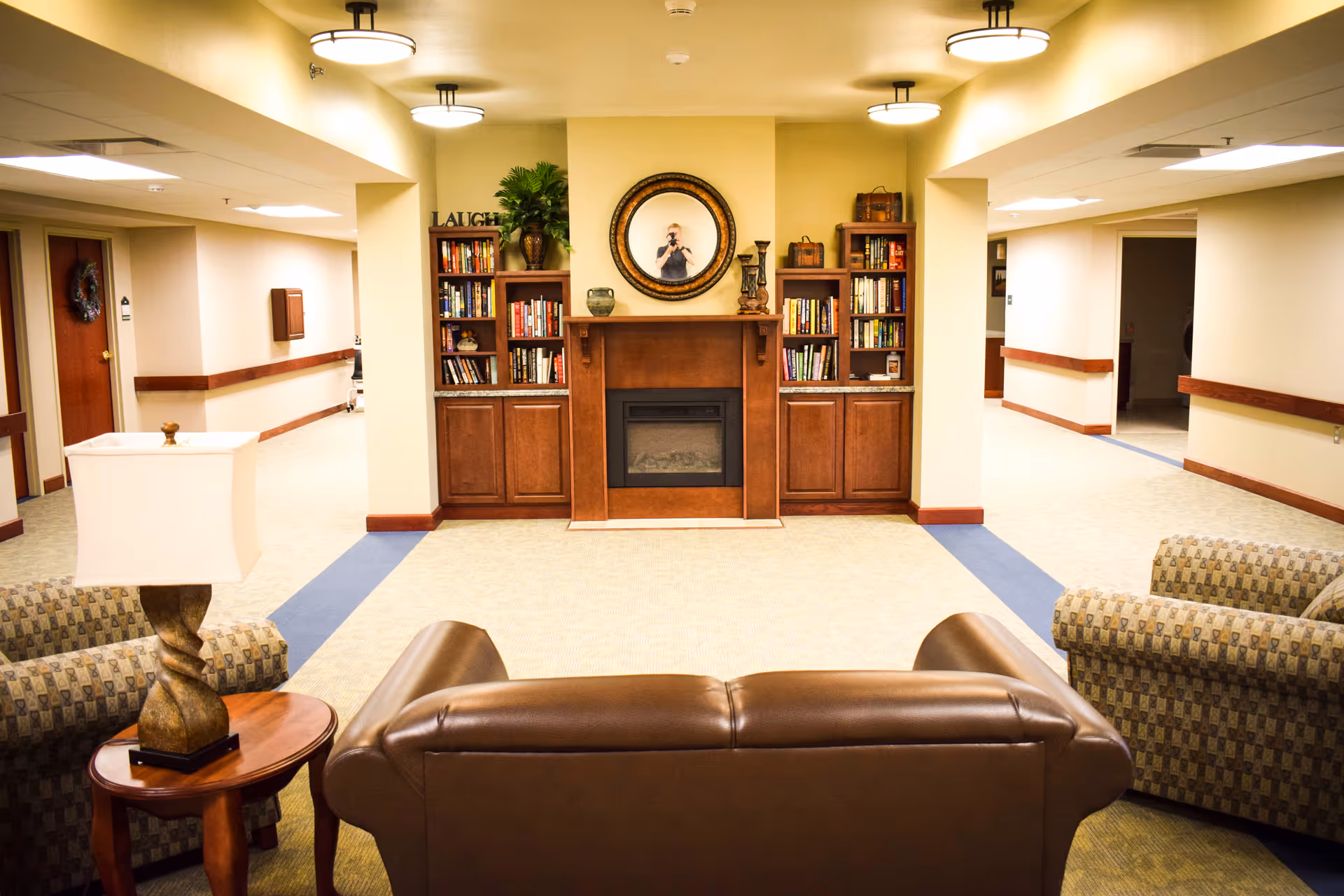 A cozy common area in a senior living facility featuring a brown leather sofa and two patterned armchairs arranged around a wooden side table with a lamp. In the background, there is a wooden fireplace flanked by built-in bookshelves filled with books and decorative items. Above the fireplace hangs a round mirror. The area is well-lit with ceiling lights and has beige walls and carpeted floors with a blue border.