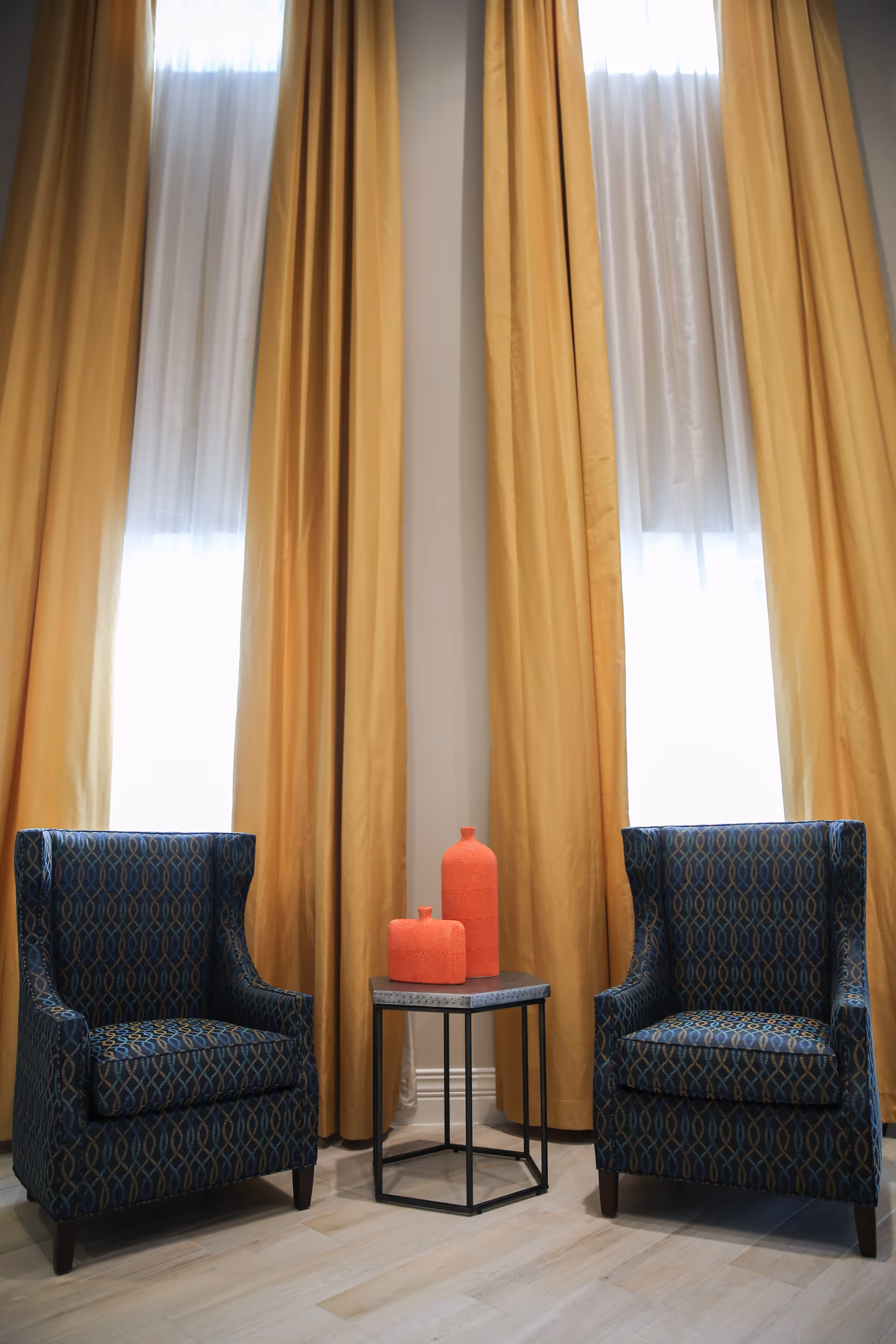 Two patterned blue armchairs with a small black metal side table between them, holding two red decorative vases. Behind the chairs are tall windows with sheer white curtains and long mustard yellow drapes.
