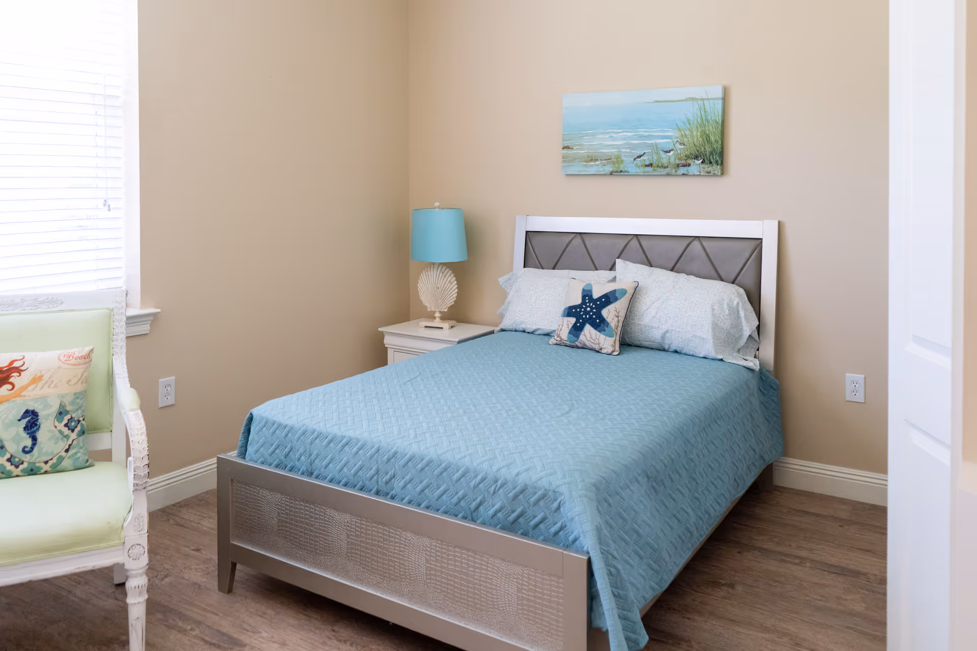A bedroom with a bed covered in a light blue quilt and matching pillows, including one with a starfish design. There is a white nightstand with a blue lamp and a seashell decoration next to the bed. A painting of a coastal scene hangs on the beige wall above the bed. A green chair with a decorative pillow featuring sea creatures is positioned near a window with white blinds.