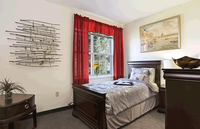 A neatly made single bed with gray floral bedding in a small bedroom. The room has a window with red curtains, a small round wooden side table with a plant, a dark wooden dresser with a decorative bowl, and a framed painting on the wall.