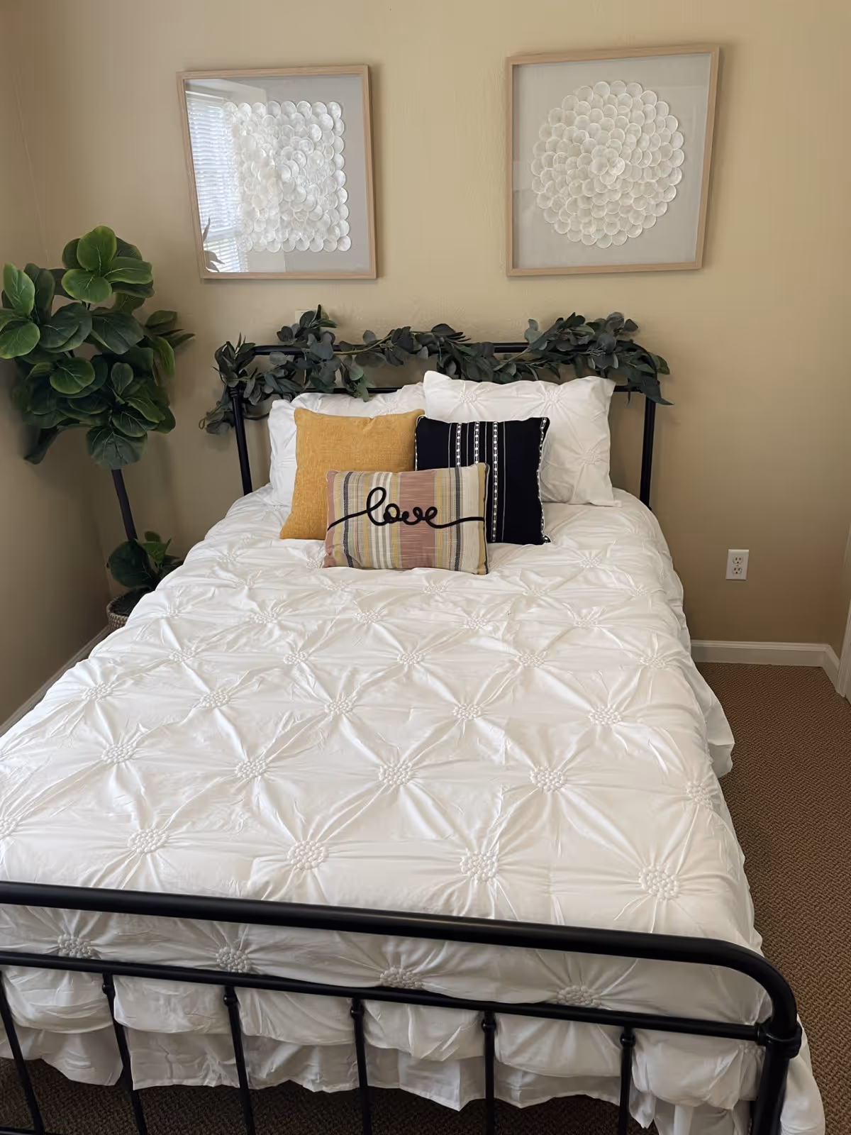 A neatly made bed with a black metal frame, white textured bedding, and three decorative pillows including one with the word 'love' on it. Above the bed are two framed floral artworks. A green leafy plant is placed in the corner of the beige-colored room.