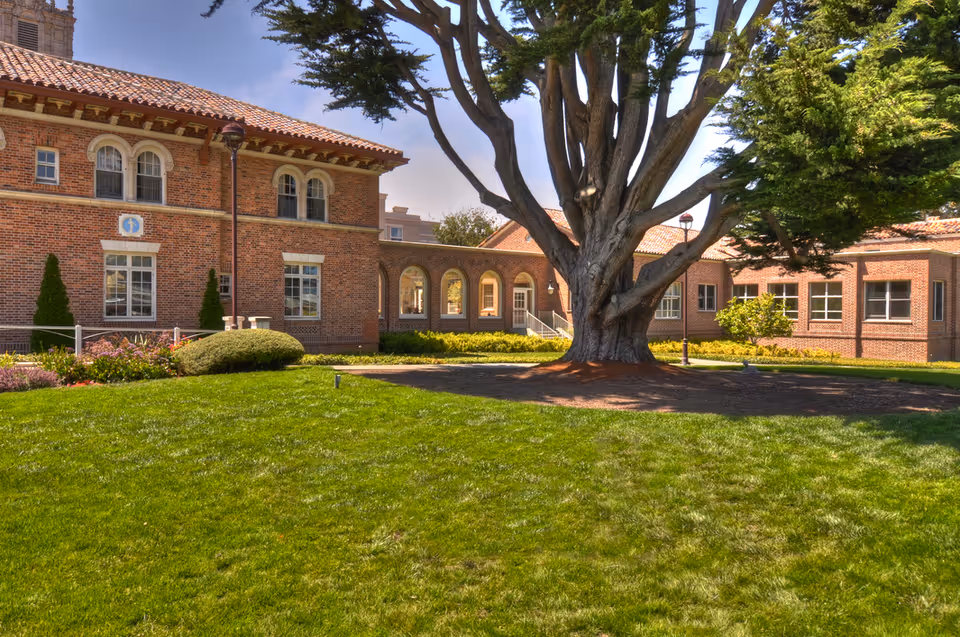 A large brick building with arched windows and a tiled roof surrounding a courtyard with a large tree in the center and well-maintained green grass and shrubs under a clear blue sky.