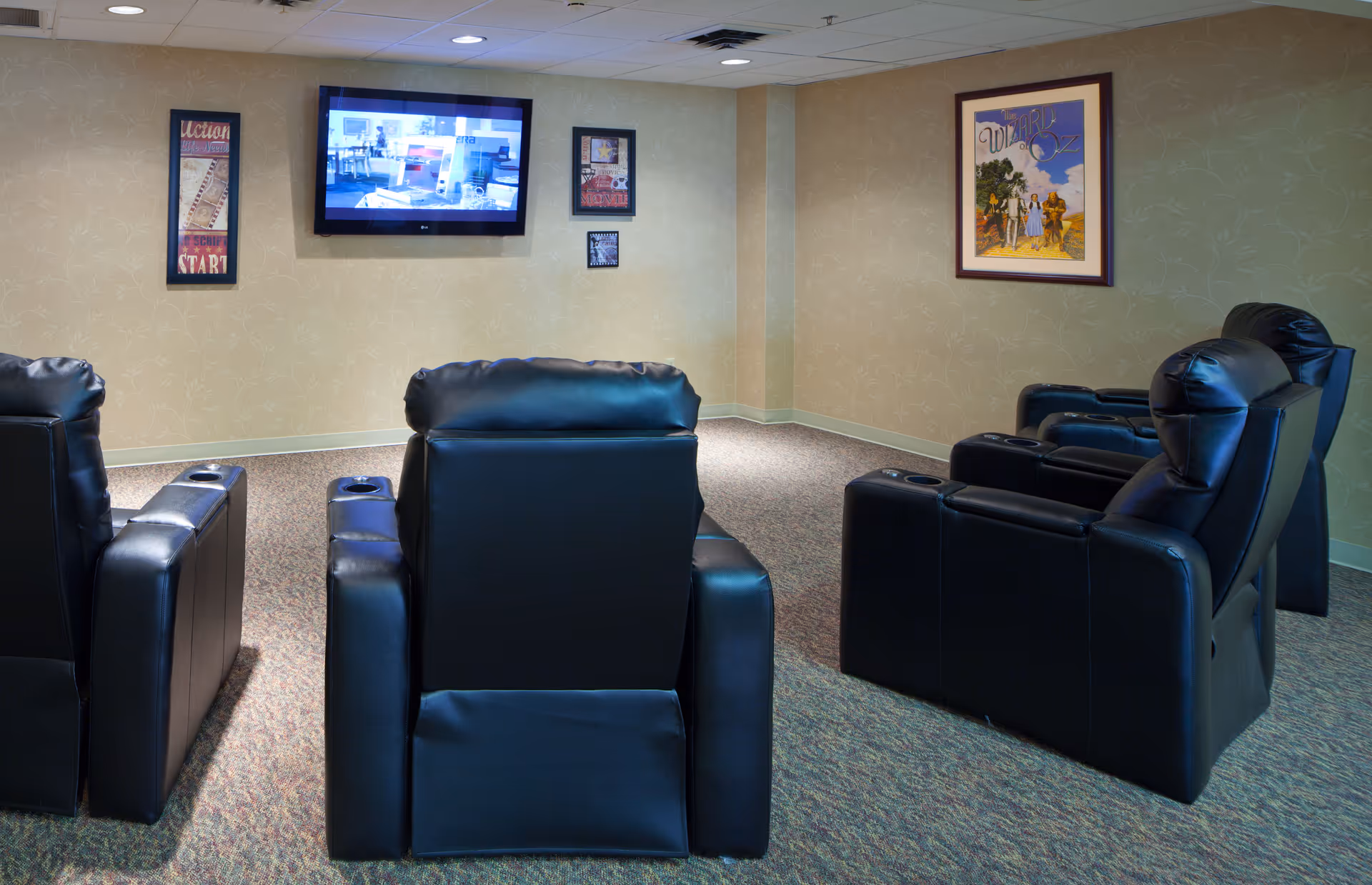 A small media room with four black leather recliners arranged in two rows facing a wall-mounted flat screen TV. The walls are beige with framed artwork, and the floor is carpeted.