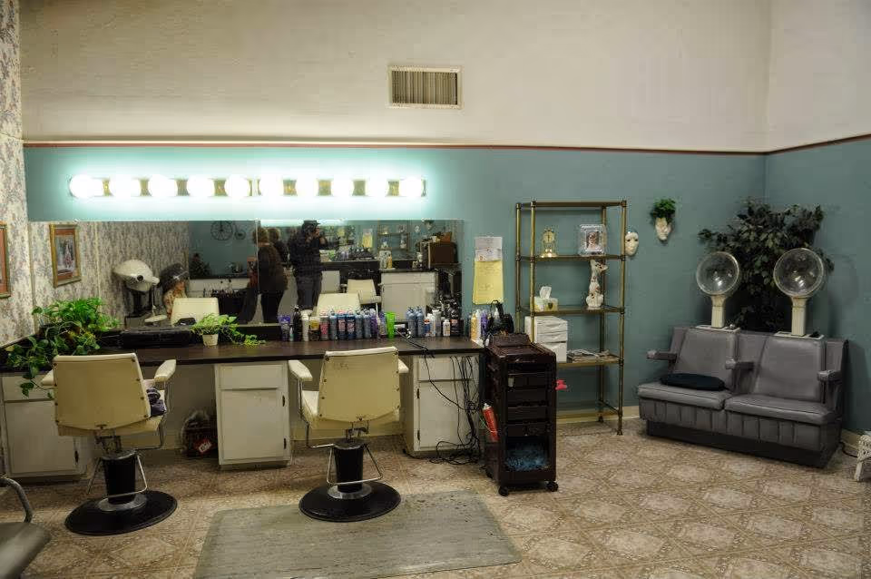 Interior view of a hair salon area with two salon chairs in front of a large mirror with bright lights above it. Various hair care products are arranged on the counter. To the right, there is a metal shelving unit with decorative items and a small couch with two vintage hair dryers. The walls are painted teal with a white upper section, and the floor has a patterned tile design.