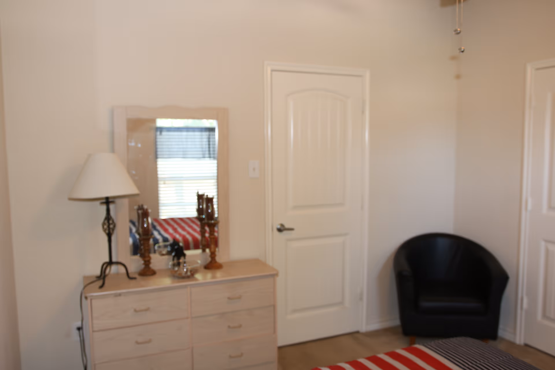 Small bedroom with a dresser and mirror, table lamp, black armchair, and a closed white door.