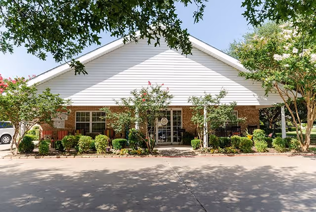 Front exterior view of a single-story building with a white gabled roof and brick walls, surrounded by small trees and shrubs, with a concrete driveway in front under a clear blue sky.