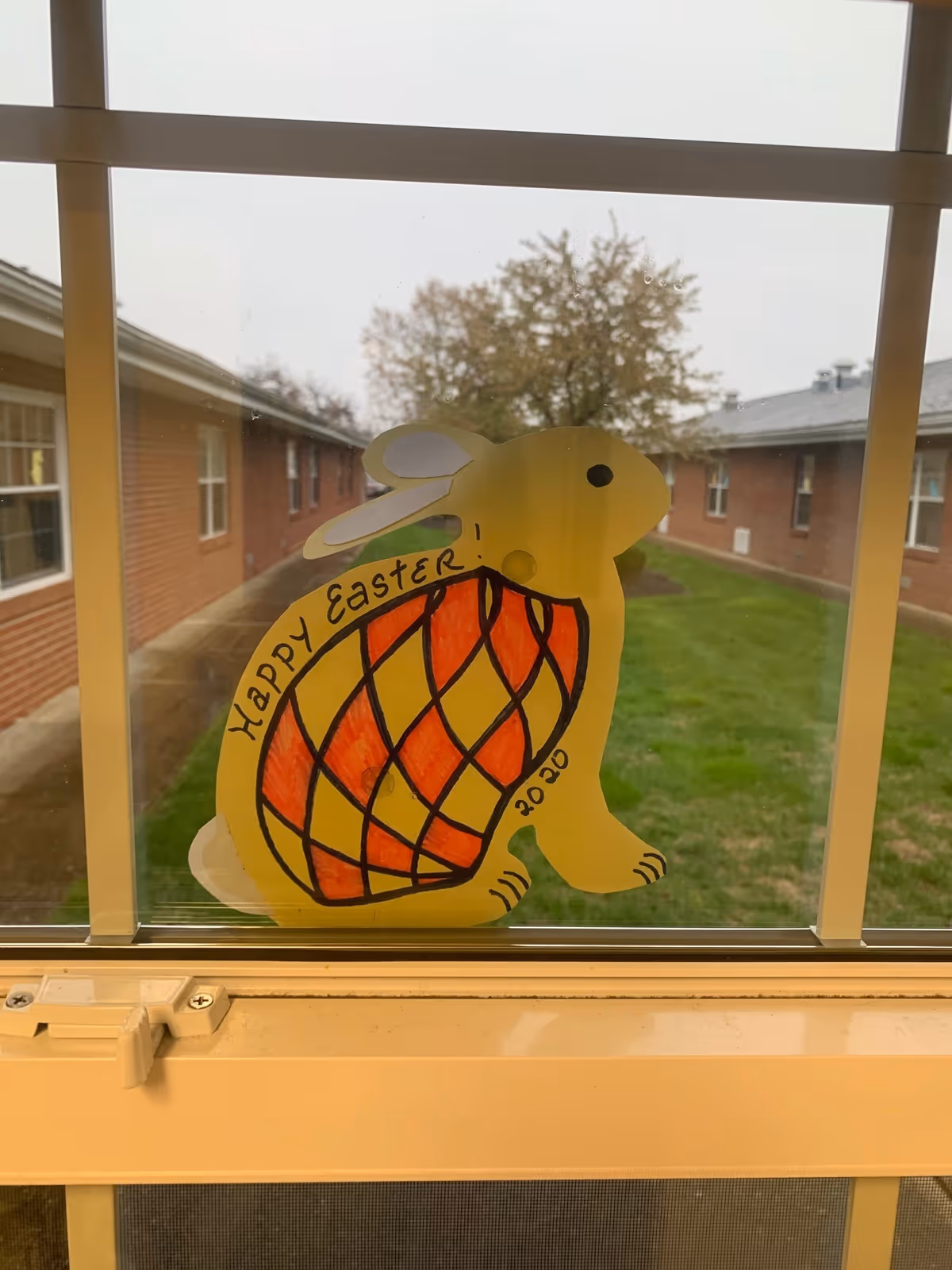 A yellow paper cutout of a rabbit with a red and yellow patterned body is taped to a window. The rabbit has the words 'Happy Easter!' written on its side along with the year '2020'. Outside the window, there is a grassy courtyard surrounded by brick buildings and trees with autumn foliage.