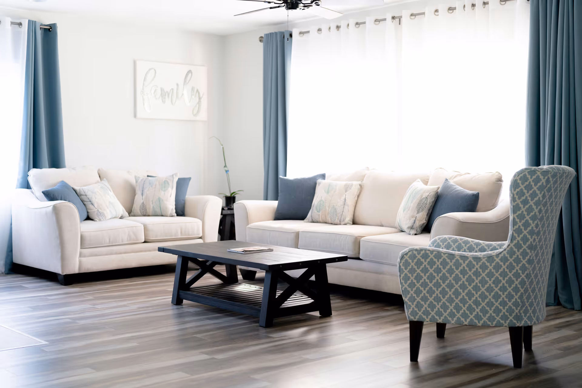 Bright living room with two beige sofas adorned with patterned and solid blue pillows, a blue patterned armchair, a dark wooden coffee table, light wood flooring, blue curtains, and a wall art piece with the word 'family'.