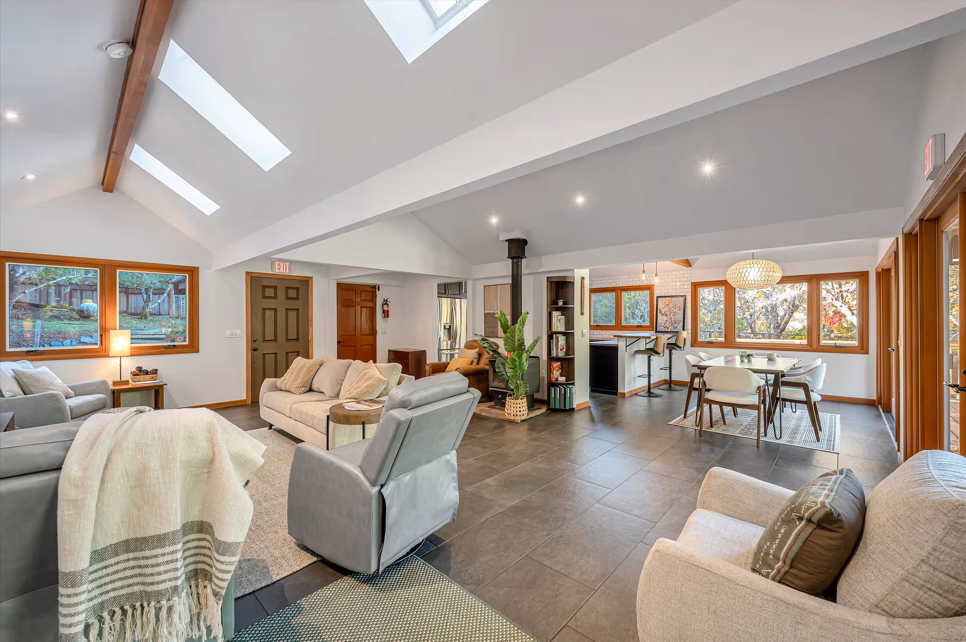 A spacious and bright senior living common area with modern furniture including sofas, armchairs, and a dining table with chairs. Large windows and skylights allow natural light to fill the room. The space features a tiled floor, a small bookshelf, and a wood stove. The walls are white with wooden trim around the windows and doors.