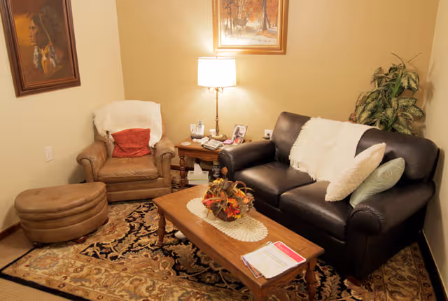 A cozy living room with a dark leather sofa adorned with white and light-colored pillows and a white throw blanket. Next to the sofa is a brown armchair with a red pillow and a white throw draped over the back. A wooden coffee table with a decorative centerpiece and some papers sits on a patterned area rug. A side table with a lamp, phone, and framed photo is positioned between the sofa and armchair. The walls are decorated with two framed paintings, and there is a green leafy plant in the corner.